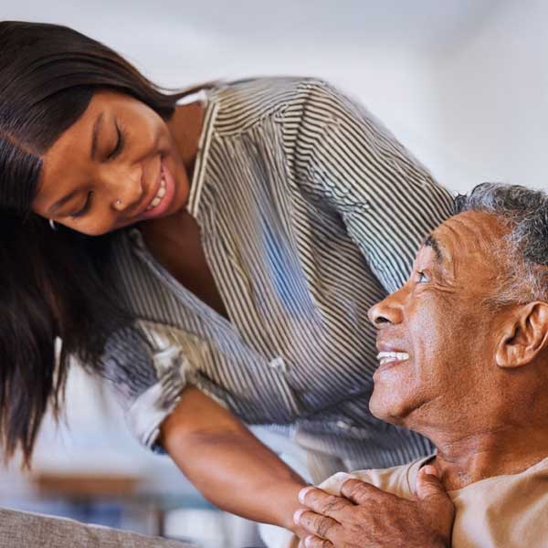 A young woman smiles warmly at an older man who is sitting down, looking up at her with a joyful expression. The woman gently holds the man's arm, showing care and affection.