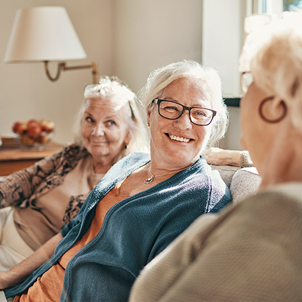 Three older women sit together on a couch, smiling and chatting in a cozy, sunlit living room. A lamp and a bowl of fruit are visible in the background, creating a warm atmosphere.