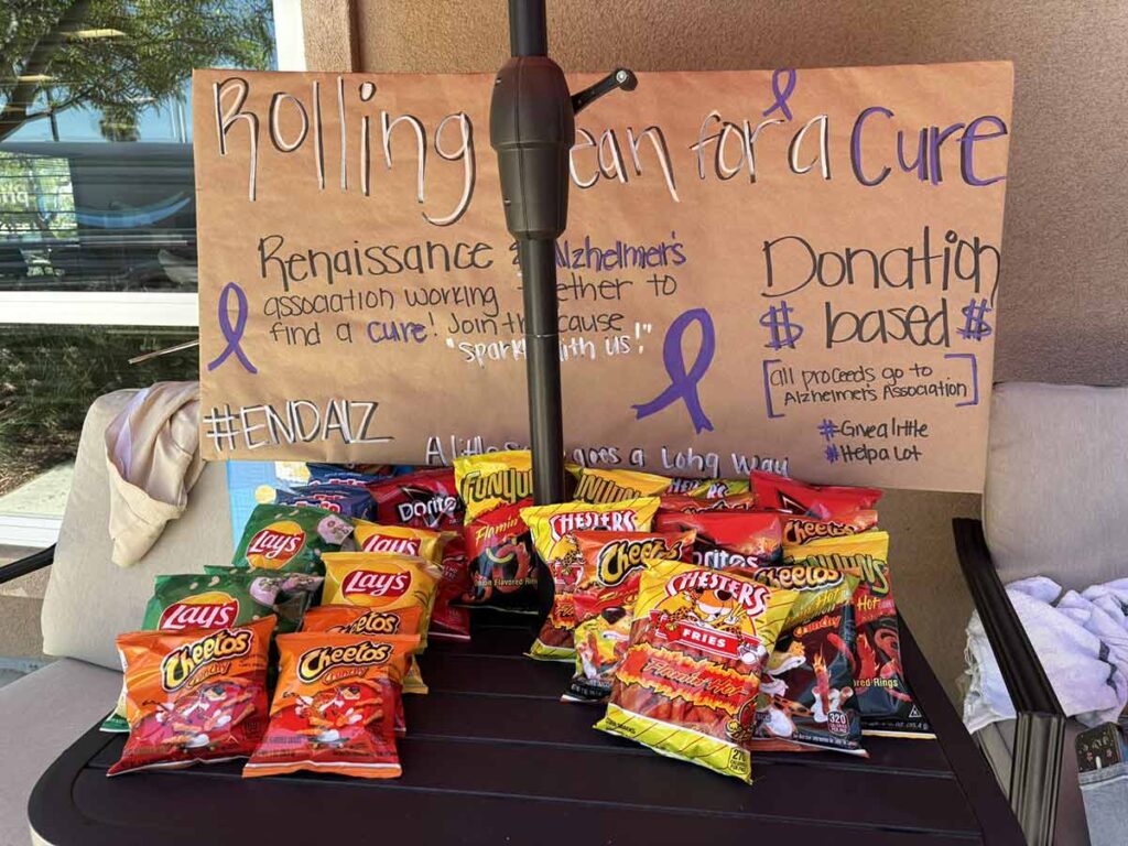 A table holds assorted chips bags in front of a sign reading “Rolling Can for a Cure.” The sign mentions supporting the Alzheimer’s Association and encourages donations to help fund a cure.