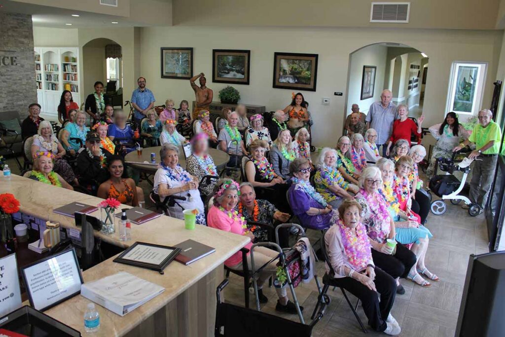 A group of elderly people, some in wheelchairs, gather in a decorated common room wearing colorful leis and Hawaiian-themed attire, smiling and posing for a group photo at what appears to be a care facility or community center.