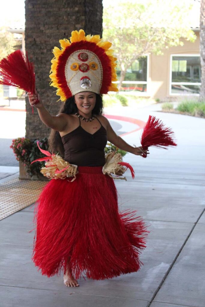 A woman wearing a red grass skirt, brown top, large decorative headdress, and holding red pom-poms, performs a traditional Polynesian dance outdoors on a covered walkway.