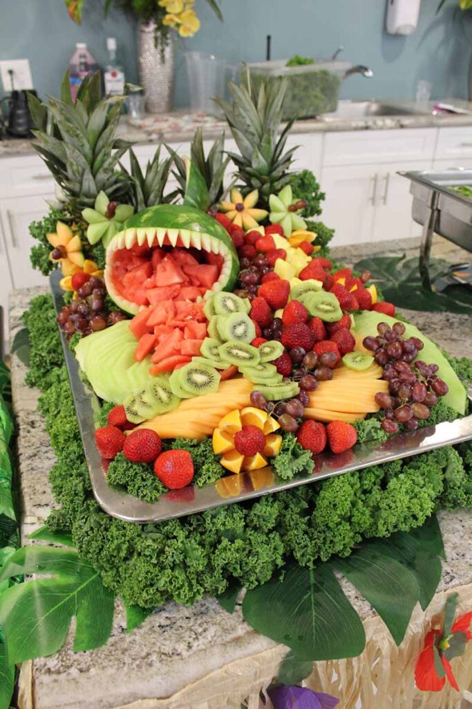 A fruit platter arranged to look like an open-mouthed monster, with a watermelon head, pineapple leaves as spikes, and an assortment of sliced fruit including strawberries, grapes, and kiwi.