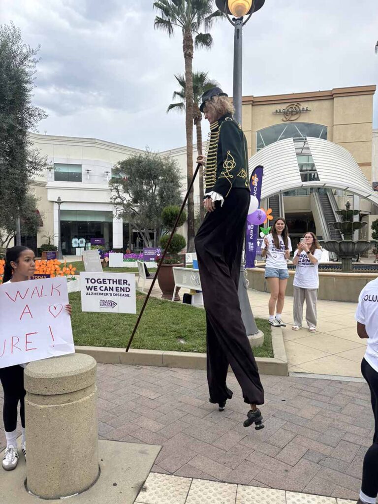 A performer on stilts in a green jacket entertains people at an outdoor event. Nearby, there are signs about ending Alzheimer’s and participants holding balloons, with shops and palm trees in the background.