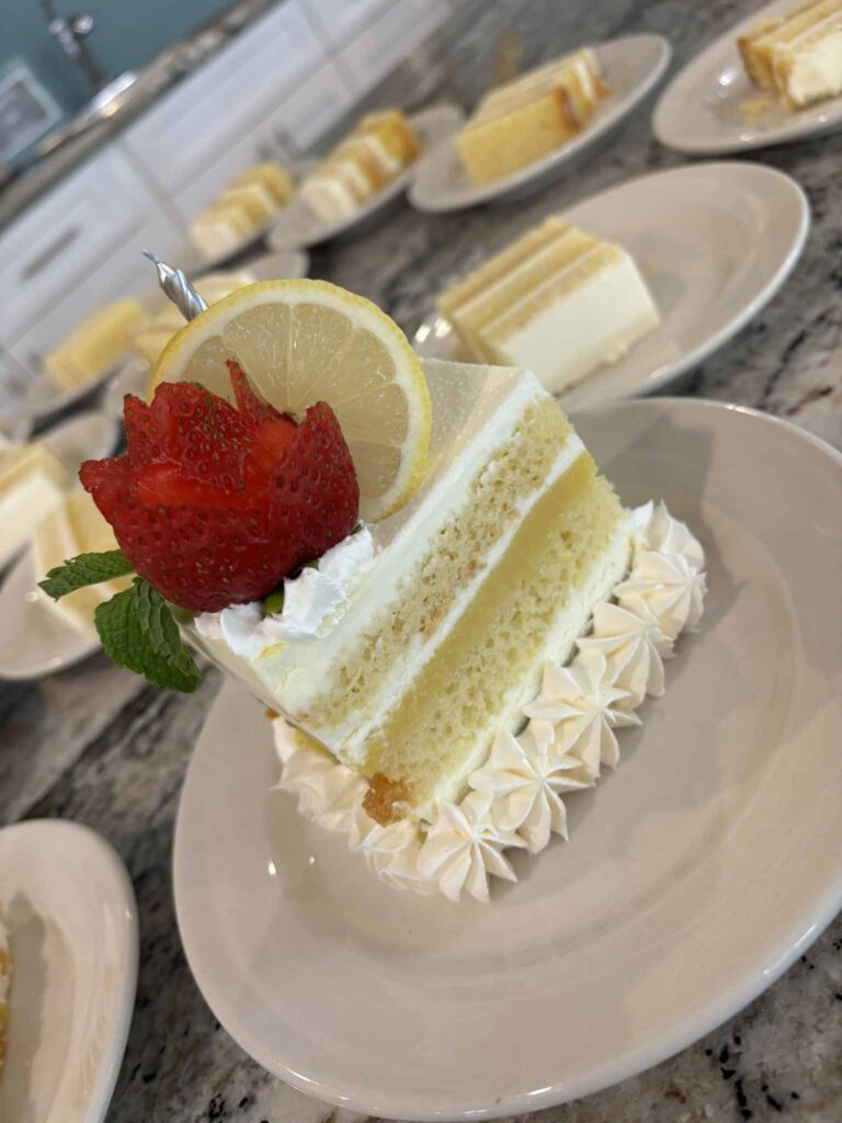 A slice of lemon cake with white frosting, topped with a fresh strawberry, a lemon slice, and a sprig of mint on a white plate. More slices of cake are visible in the background on similar plates.