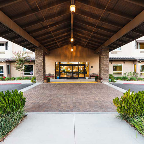 A covered entrance with stone pillars leads to double glass doors at a building, with brick paving, plants, and exterior lights illuminating the entryway.
