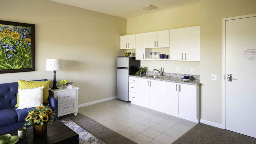 A small kitchen with white cabinets, a granite countertop, a stainless steel fridge, and a sink. To the left, a blue sofa, side table with a lamp, and sunflowers. A colorful flower painting hangs on the wall above the sofa.