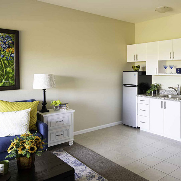 A small, modern studio apartment kitchen with white cabinets, stainless steel fridge, a blue sofa with colorful pillows, a white side table, sunflowers on a coffee table, and a floral painting on the wall.
