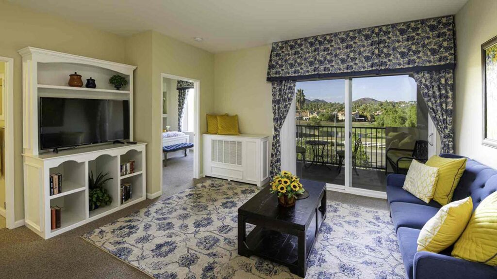 Bright living room with a blue couch, yellow pillows, a coffee table with sunflowers, TV on a white stand, and large window with blue curtains overlooking a balcony and scenic outdoor view.