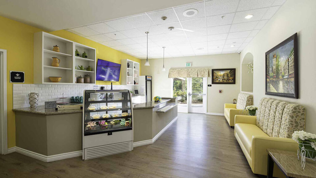 A brightly lit café with yellow walls, a display case of desserts, white shelves with pottery, cushioned yellow seating, potted flowers, a TV, and a large window letting in natural light.