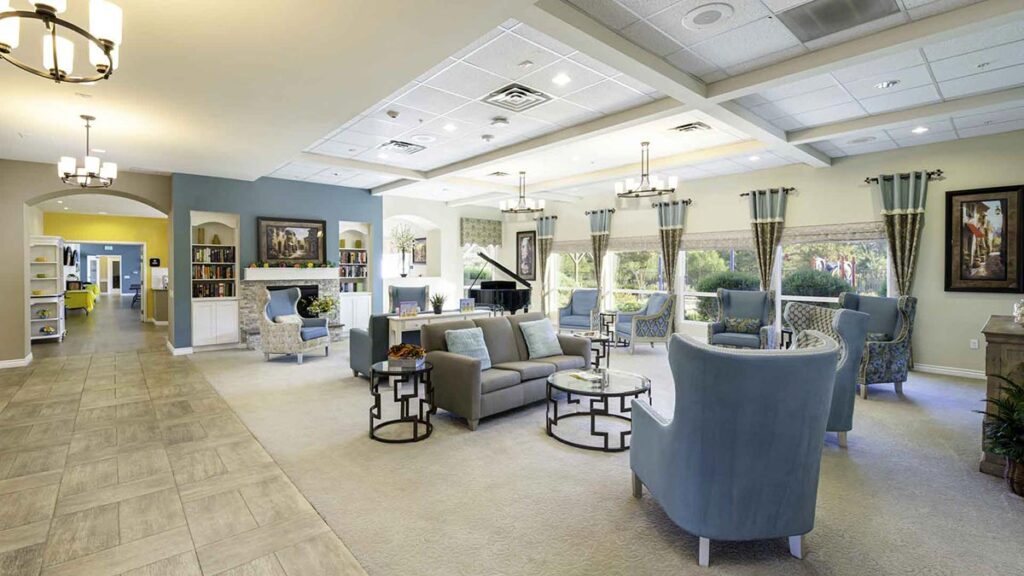 Spacious, elegant living room with modern armchairs, sofas, coffee tables, large windows, a fireplace, chandeliers, a piano, and decorative art. A hallway with bookshelves is visible to the left.