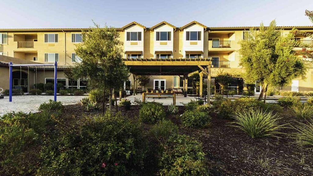 A three-story apartment building with tan walls and multiple windows, seen behind a landscaped garden with shrubs, small trees, and a pergola-covered seating area in the courtyard.