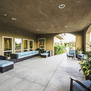 Covered outdoor patio with cushioned wicker seating along the walls, large windows on one side, and potted plants by the entrance; bright daylight and greenery visible outside.