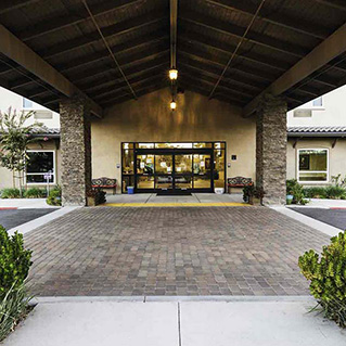 Covered entrance with stone pillars leading to glass doors of a building, flanked by benches and landscaped greenery, with warm lighting under the roof.
