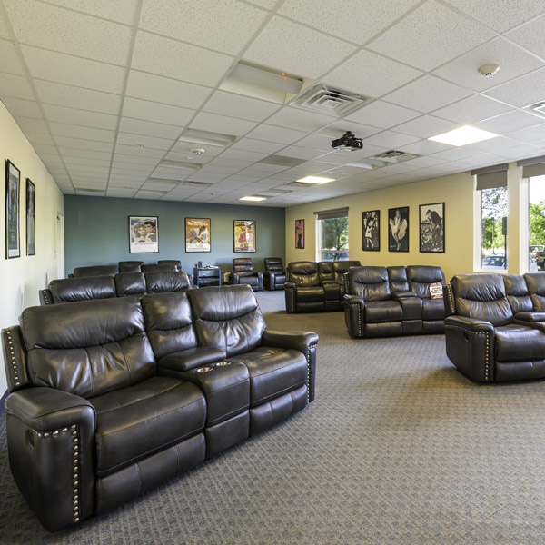 Room with several black leather recliner sofas arranged in rows, movie posters on the walls, carpeted floor, and large windows letting in natural light. Appears to be a home theater or media/lounge room.