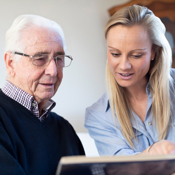 An elderly man with glasses sits next to a younger woman with blonde hair. They are looking at a photo album together, smiling as the woman points to something on the page.
