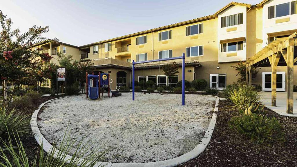 A small playground with swings and play equipment sits in a sandy area, surrounded by plants and shrubs, in front of a beige three-story apartment building with multiple windows and balconies.