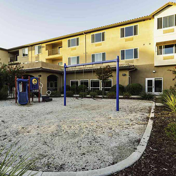 A playground with a slide and swings sits in front of a three-story beige apartment building, surrounded by shrubs and trees, under a clear sky.