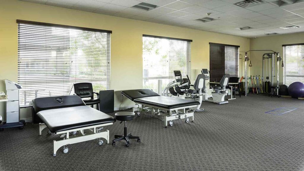 A spacious physical therapy room with large windows, treatment tables, exercise equipment, chairs, and various medical devices, all arranged neatly on a carpeted floor.