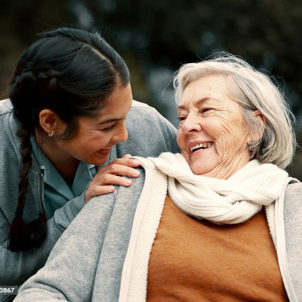 A young woman smiles warmly while touching the shoulder of an older woman in a wheelchair. Both are outdoors, sharing a joyful moment. The older woman wears a scarf and cardigan, and both appear happy and connected.