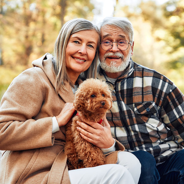 Smiling older couple sitting outdoors, the woman in a tan coat, the man in a plaid jacket, both holding a small curly-haired brown dog, with trees and soft sunlight in the background.