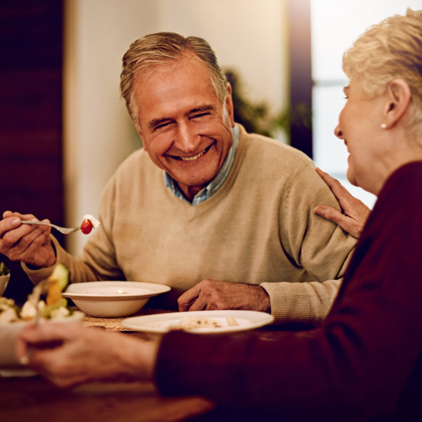 An older man and woman are sitting at a table, smiling and talking while enjoying a meal together. The man is holding a spoon and appears to be laughing, creating a warm, joyful atmosphere.