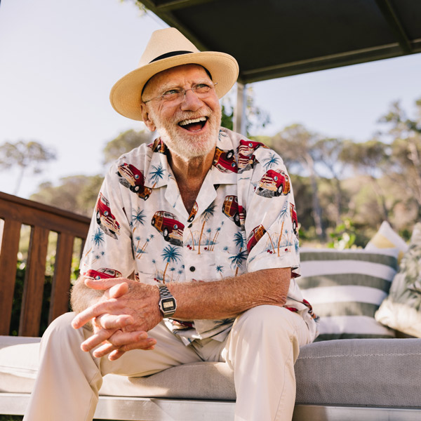 An elderly man in a straw hat and colorful shirt with vans and palm trees sits outdoors on a sofa, smiling and laughing on a sunny day.