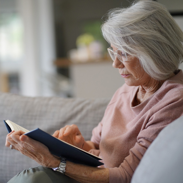 An older woman with gray hair and glasses sits on a sofa, wearing a pink sweater and reading a book. She appears focused, with one hand holding the book and the other pointing at the page.