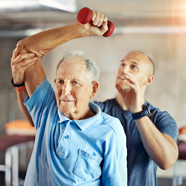 An older man in a light blue polo shirt lifts a small red dumbbell with his right hand, assisted by a younger trainer in a gym or rehabilitation setting.