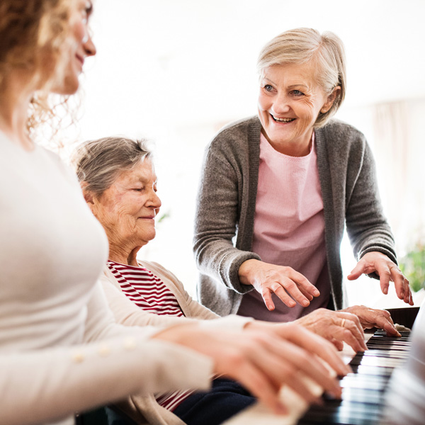 Three women, spanning different ages, are smiling and playing the piano together. The older woman in a striped shirt sits at the keys, while the others stand nearby, enjoying the moment. The scene is bright and cheerful.