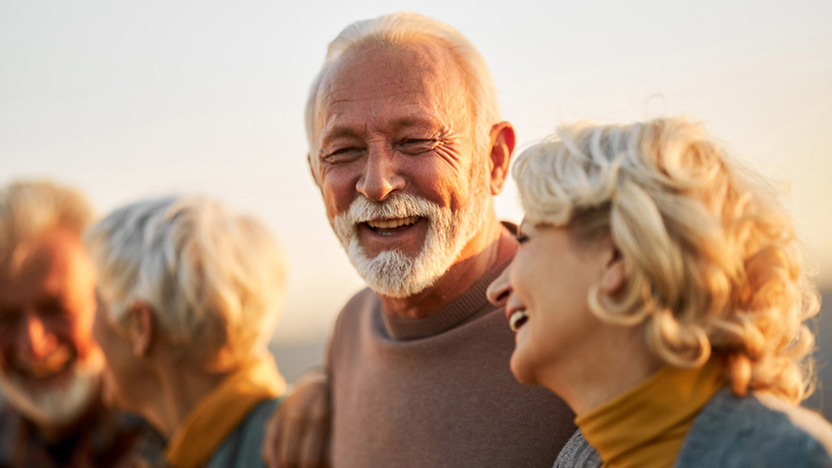 Four older adults with gray hair are outdoors, laughing and smiling together in warm, natural sunlight. The focus is on a bearded man and a woman next to him, both appearing happy and relaxed while enjoying the best level of care.
