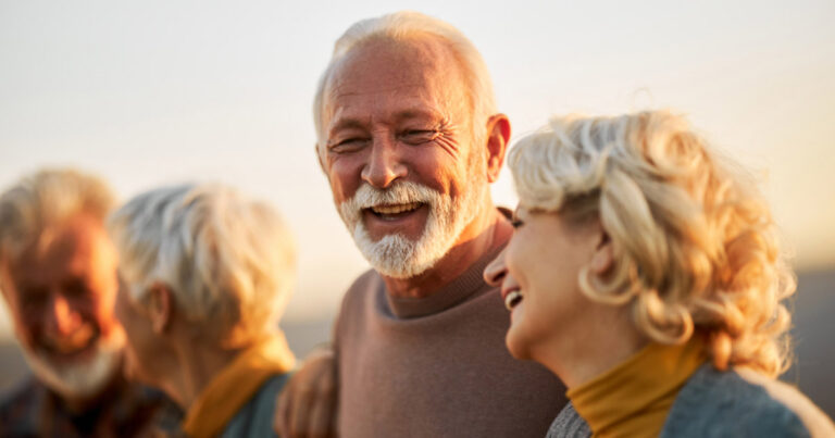 A group of older adults with gray hair and beards are smiling and laughing together outdoors, enjoying each other's company in warm, natural sunlight.