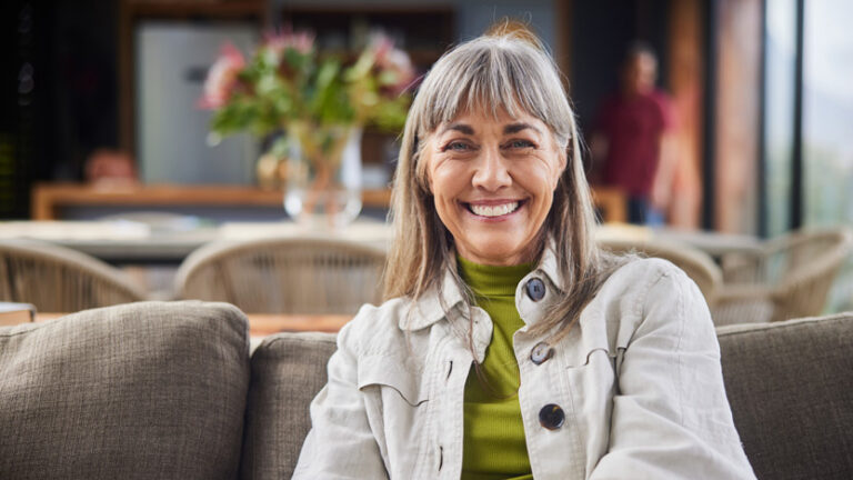 An older woman with gray hair and bangs is smiling while sitting on a couch indoors, enjoying the best level of care. She is wearing a light-colored jacket over a green turtleneck, with a blurred room featuring a table, chairs, and flowers in the background.