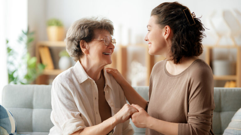 An elderly woman and a young woman sit on a couch, smiling and holding hands, sharing a warm, happy moment together in a bright, cozy living room—a perfect example of the connections fostered in supportive living.